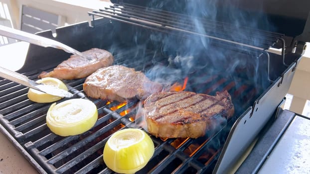 Ribeye steaks are sizzling alongside golden grilled onions on a barbecue grill, with wisps of smoke hinting at the flavorful feast being prepared.