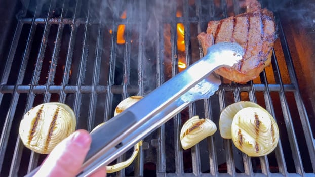 Ribeye steaks are sizzling alongside golden grilled onions on a barbecue grill, with wisps of smoke hinting at the flavorful feast being prepared.