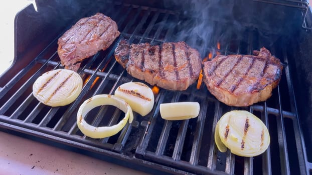 Ribeye steaks are sizzling alongside golden grilled onions on a barbecue grill, with wisps of smoke hinting at the flavorful feast being prepared.