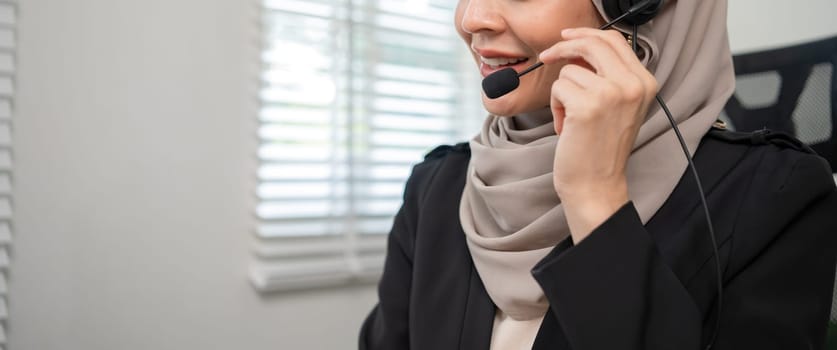 Call center worker, young Muslim woman wearing hijab, talking to customer on call phone on computer in customer service office.