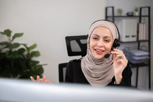 Call center worker, young Muslim woman wearing hijab, talking to customer on call phone on computer in customer service office.