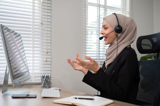 Call center worker, young Muslim woman wearing hijab, talking to customer on call phone on computer in customer service office.