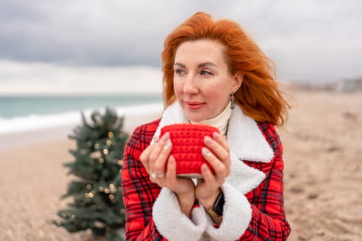 Lady in plaid shirt with a red mug in her hands enjoys beach with Christmas tree. Coastal area. Christmas, New Year holidays concep.