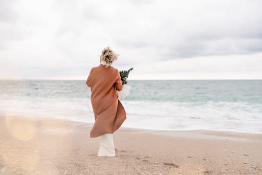 Blond woman Christmas sea. Christmas portrait of a happy woman walking along the beach and holding a Christmas tree in her hands. She is wearing a brown coat and a white suit