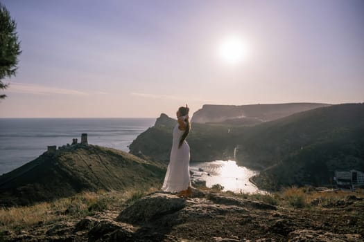 woman stands on a rocky hill overlooking a body of water. She is wearing a white dress and she is enjoying the view