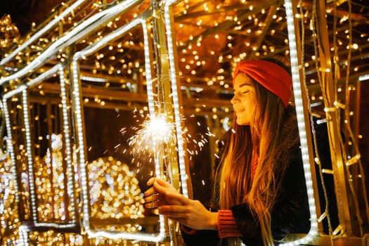 Woman holding sparkler night while celebrating Christmas outside. Dressed in a fur coat and a red headband. Blurred christmas decorations in the background. Selective focus.