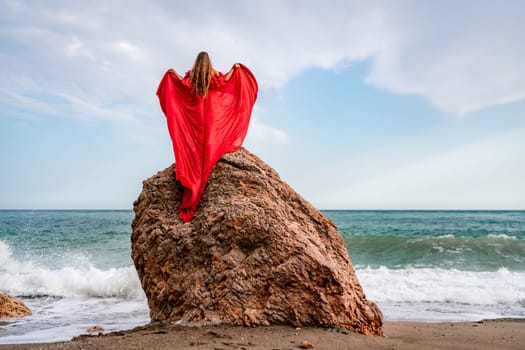 woman sea red dress. Woman with long hair on a sunny seashore in a red flowing dress, back view, silk fabric waving in the wind. Against the backdrop of the blue sky and mountains on the seashore