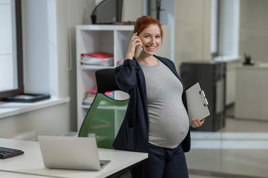 Pregnant woman using mobile phone and holding paper tablet in office