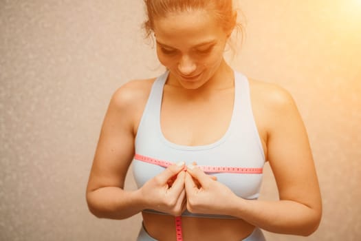 Cropped view of slim woman measuring breasts with tape measure at home, close up. A European woman checks the result of a weight loss diet or liposuction indoors. Healthy lifestyle