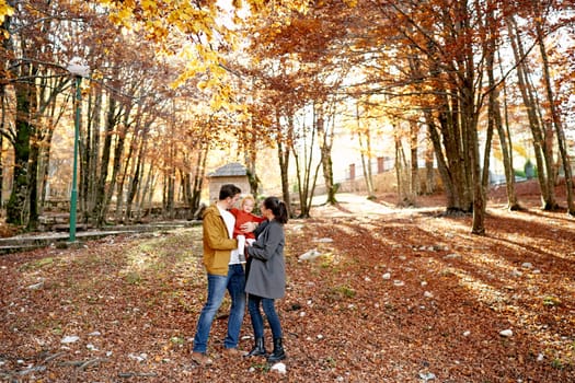 Mom and dad hold a little girl in their arms and look at her while standing in the autumn park. High quality photo