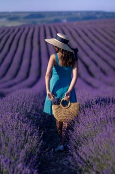 A woman wearing a straw hat and a blue dress is walking through a field of lavender. She is holding a basket and she is enjoying the scenery