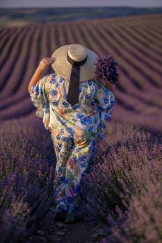 A woman wearing a straw hat and a floral dress walks through a field of lavender. She is holding a bouquet of flowers in her hand