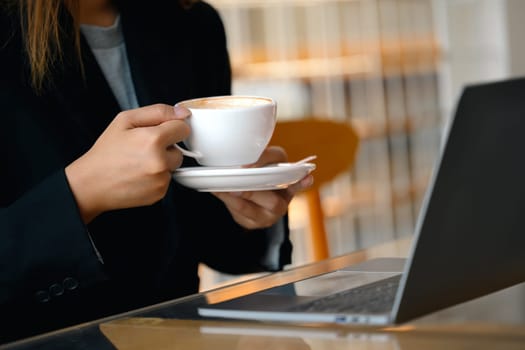 Businesswoman holding a cup of coffee sitting with laptop in modern cafe.