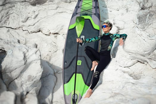 beautiful sup surfer girl in a wetsuit on the ocean shore against a beautiful blue sky poses sexually with a paddle