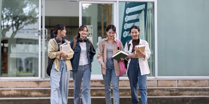 Four young women are walking down a sidewalk, each carrying a book. They are dressed in casual clothing and appear to be students. Scene is relaxed and friendly