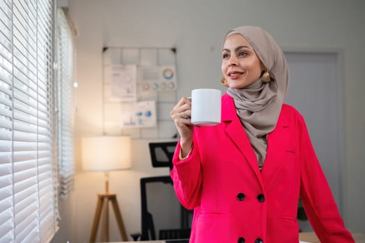 Confident Muslim businesswoman standing with coffee mug in front of desk in office during morning time.