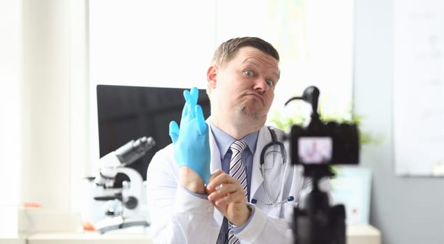 Portrait of joyful doctor practitioner wearing blue sterile glove on hands and making strange face to high-tech camera in modern hospital office. Healthcare concept