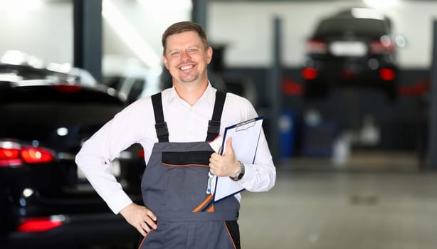 Portrait of smiling man holding paper folder with information about different autos and standing in modern car maintenance garage. Machinery repairman concept. Blurred background