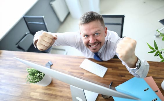 Top view of cheerful businessman standing near computer and looking at camera with happiness. Worker winning prize or signing profitable contract. Blurred background