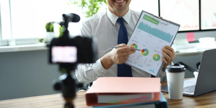 Focus on financial documents with statistics data in male hands. Blogger showing stats and pointing with pencil to important diagram for camera. Man recording video for vlog. Blurred background