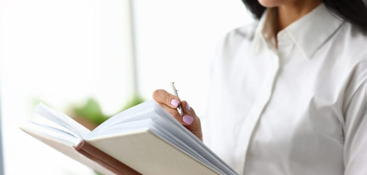 Focus on arms of smart businesswoman writing something in paper tablet with metallic pen. Classy woman wearing fashionable outfit in modern office. Accounting office concept.