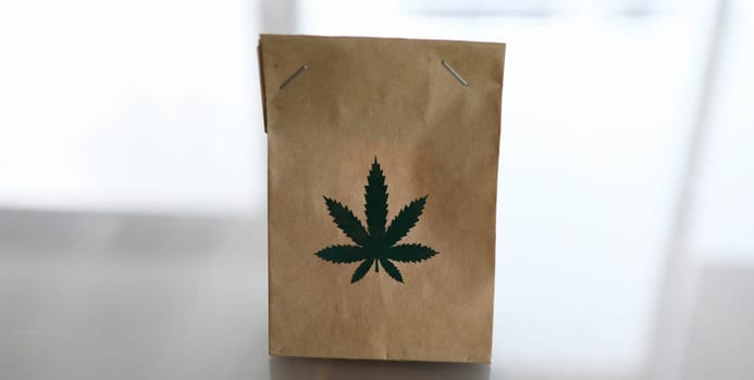 Bag of kraft brown paper with the marijuana logo lies on the table closeup background