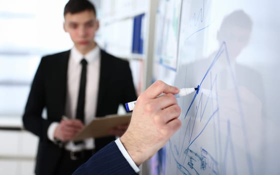 Focus on male hand writing growth income on desk with special pen. Attractive employee in classy suit with tie looking at boss with concentration. Company meeting concept. Blurred background