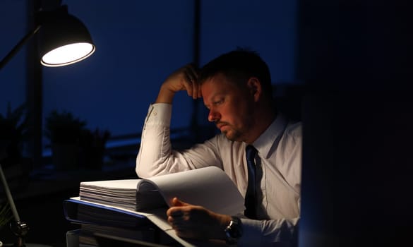 Portrait of stressed businessman holding important documents and reading corporate information. Man holding hand near aching forehead. Night at office concept