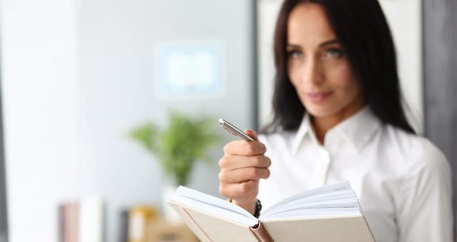 Focus on hardworking businesswoman hand holding metallic pen and note book. Smart woman standing in modern office and looking somewhere. Successful job concept