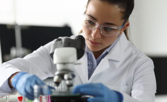 Adult caucasian beauty woman chemist in protective glasses looking at microscope against chemistry lab background. Medical analysis processing concept