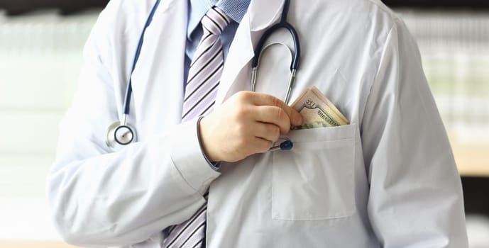 Focus on male hands holding green banknotes and putting big amount of money in pocket of white uniform with stethoscope. Healthcare and clinic concept