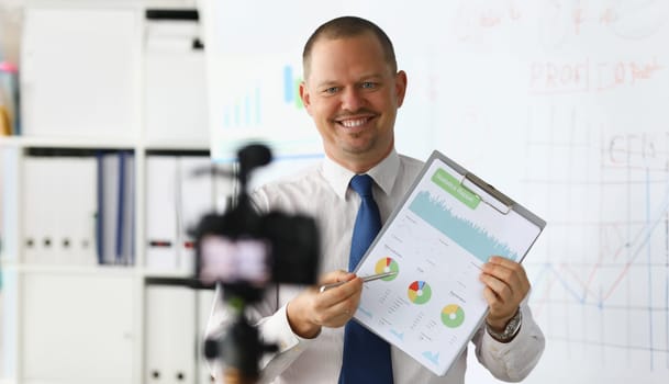 Portrait of smiling blogger in classic suit and tie showing statistics data with graphs charts diagram. Promo videoblog or photo session in office camcorder to tripod. Blurred background