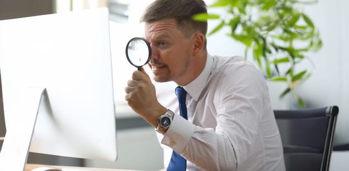 Portrait of businessman in office at computer. Manager in classic shirt and tie looking at pc monitor through magnifier attentively. Blurred background