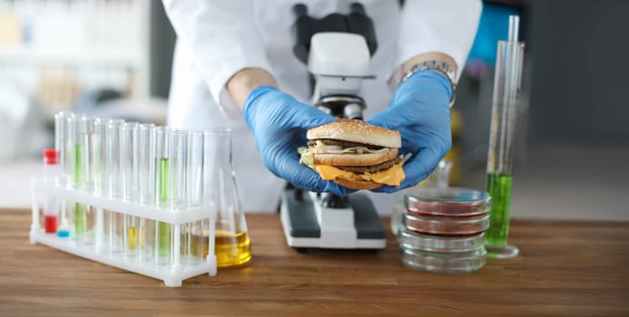 Male hand hold tasty burger in hand with blue protective gloves closeup. Testing food nutrition concept.