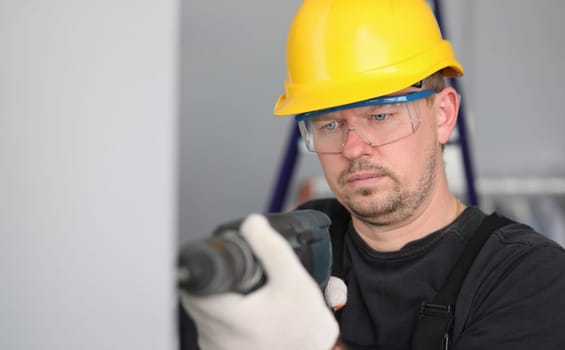 Male adult worker in yellow helmet with protective glasses hold electric drill in hand. Flat repair concept