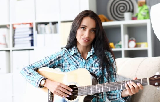 Portrait of beautiful woman playing guitar in studio. Wonderful lady wearing fashionable checkered shirt and smart clock. Music concept. Blurred background