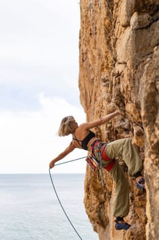 A woman is climbing a rock wall with a rope. The rope is attached to her harness and is being pulled by a person on the ground. The woman is wearing a black tank top and green pants
