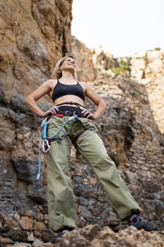 A woman in a green pair of pants stands on a rocky cliff. She is wearing a black tank top and a black harness