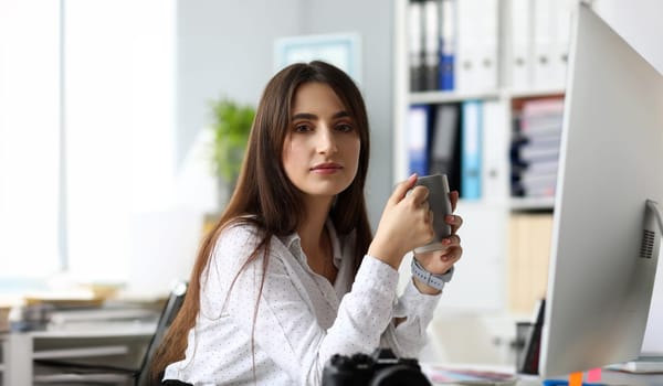 Pretty adult female sitting at worktable drinking hot beverage and looking in camera headshot