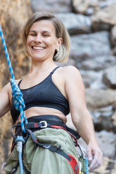 A woman is smiling while wearing a black bra and green pants. She is holding a rope and she is climbing a rock wall