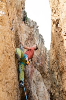 A man is climbing a rock wall with a blue rope. The man is wearing a red shirt and green pants