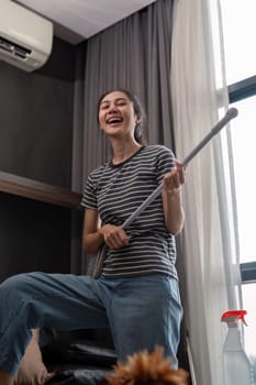 House cleaning with fun. Happy young asian housewife singing song during cleanup, using mop as guitar, enjoying domestic work. Young woman dancing and cleaning in living room.