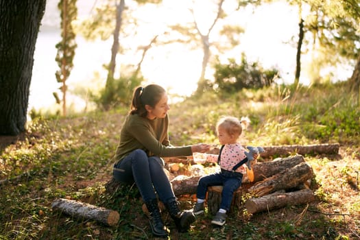 Mom feeds a little girl with porridge from a spoon sitting on stumps in the forest. High quality photo