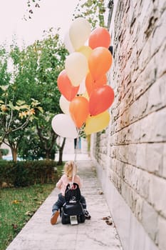 Little girl rides a toy car with tied balloons in the garden. Back view. High quality photo
