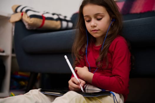 Authentic portrait little student girl in red shirt, using a white stylus, drawing on digital tablet at home. Online education and distance learning concept. Smart kid having fun on electronic device