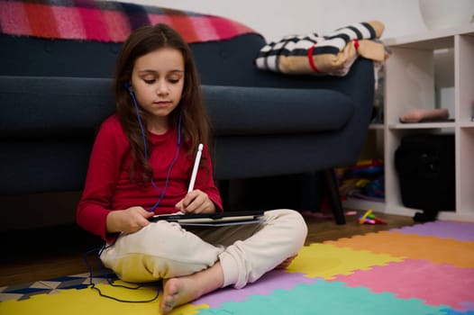 Confident primary school girl with a digital tablet at home, sitting on a colorful puzzle carpet in her kids room. Cute child focused on drawing picture on electronic device. writing using a stylus