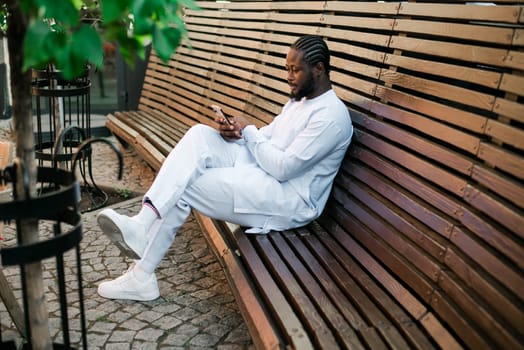 African American hipster guy in cultural and ethnic clothing dashiki chatting on smartphone outdoors, texting with friends on social networks. Millennial and Internet concept.