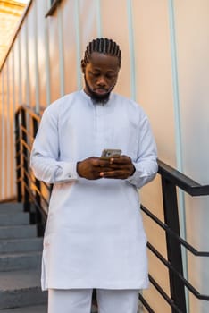 African american man in stylish ethnic dashiki wear viewing photos in social networks on mobile enjoying free time outdoors. Dark-skinned man watching funny video on smartphone and social networks. Blog and blogger scrolling content in Internet.