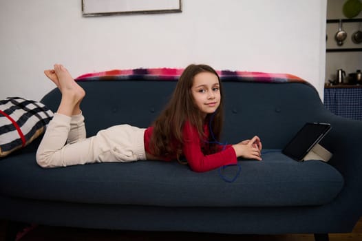 Caucasian adorable elementary age school girl smiling looking at camera, lying on sofa, browsing websites, watching movies and having fun on her digital tablet with black mockup screen.