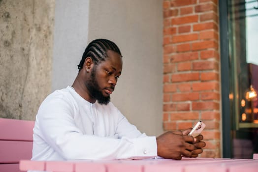 African American hipster guy in cultural and ethnic clothing dashiki chatting on smartphone outdoors, texting with friends on social networks. Millennial and Internet concept.